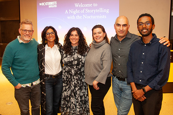 The five storytellers are posing in a line with the arms around one another in front of a welcome screen for the event